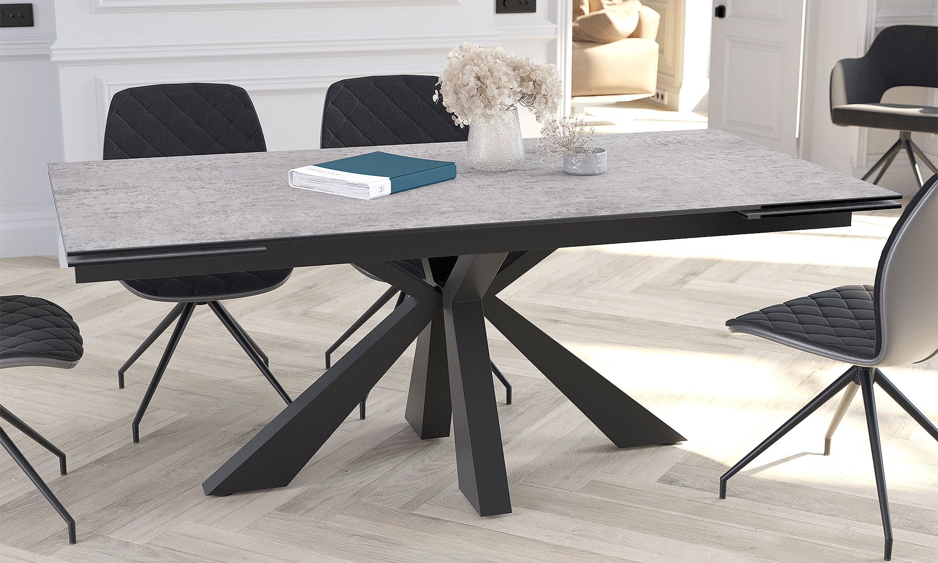 Photo d'ambiance mettant en scène une table de repas rectangulaire avec un pied centrale en étoile en acier laqué noir matte et un plateau en céramique effet béton gris. Il y a aussi des chaises noir pivotantes.